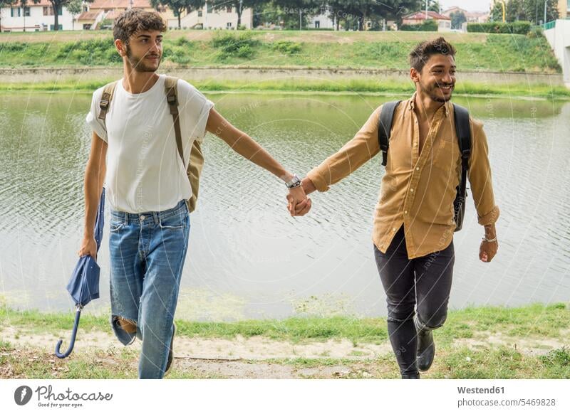 Porträt von jungen Homosexuell Paar mit Rucksäcken zu Fuß Hand in Hand am Ufer des Flusses Flussufer Rucksack gehen gehend geht schwul Homosexueller Mann