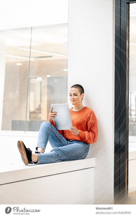 Entspannte Geschäftsfrau sitzt auf der Fensterbank eines Bürogebäudes und benutzt ein digitales Tablet Leute Menschen People Person Personen Europäisch