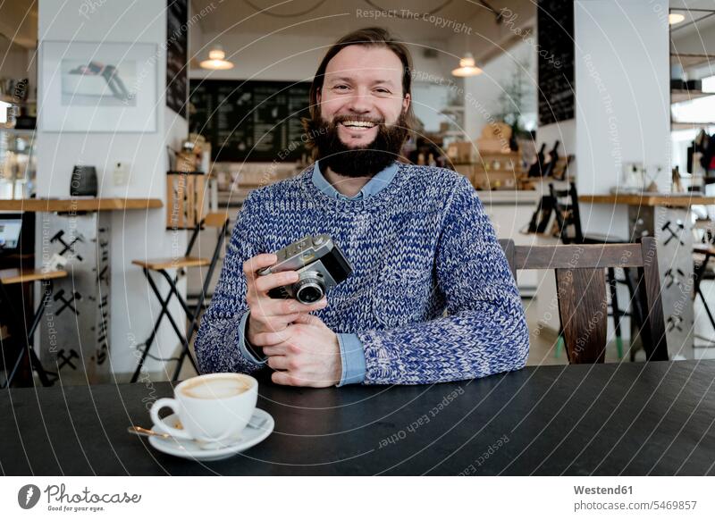Mann mit Bart sitzt im Café und hält eine alte Kamera Männer männlich Fotoapparat Fotokamera Fotograf Fotografen Photographen Vollbart Vollbärte Bärte trinken