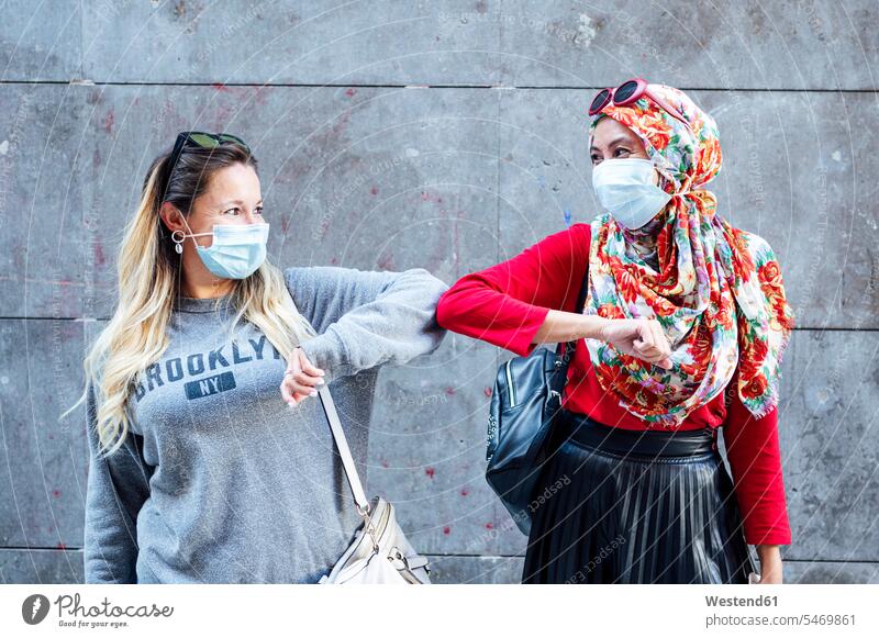 Freundinnen, die während COVID-19 Schutzmasken tragen und mit Ellbogenschlägen grüßen Farbaufnahme Farbe Farbfoto Farbphoto Außenaufnahme außen draußen
