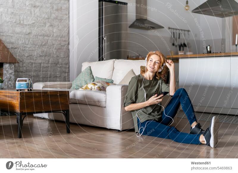Junge Frau sitzt zu Hause auf dem Boden, benutzt ein Smartphone und trägt Kopfhörer Kopfhoerer Musik hören iPhone Smartphones attraktiv schoen gut aussehend