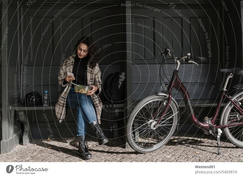 Frau mit Fahrrad beim Mittagessen und mit Smartphone auf dem Bahnsteig, Berlin, Deutschland Informationen Bänke Sitzbank Sitzbänke Transport Transportwesen