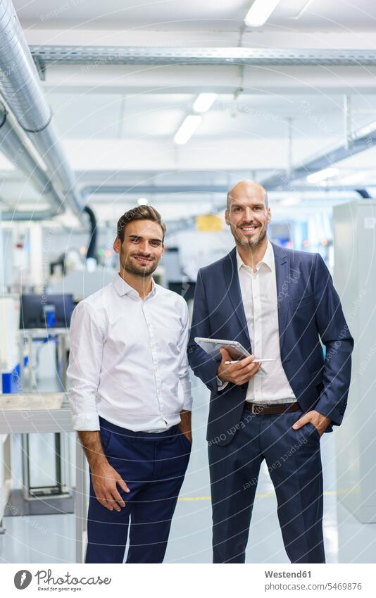 Lächelnder reifer Geschäftsmann hält digitales Tablett in der Hand, während er einem jungen Ingenieur in der Fabrik zur Seite steht Farbaufnahme Farbe Farbfoto