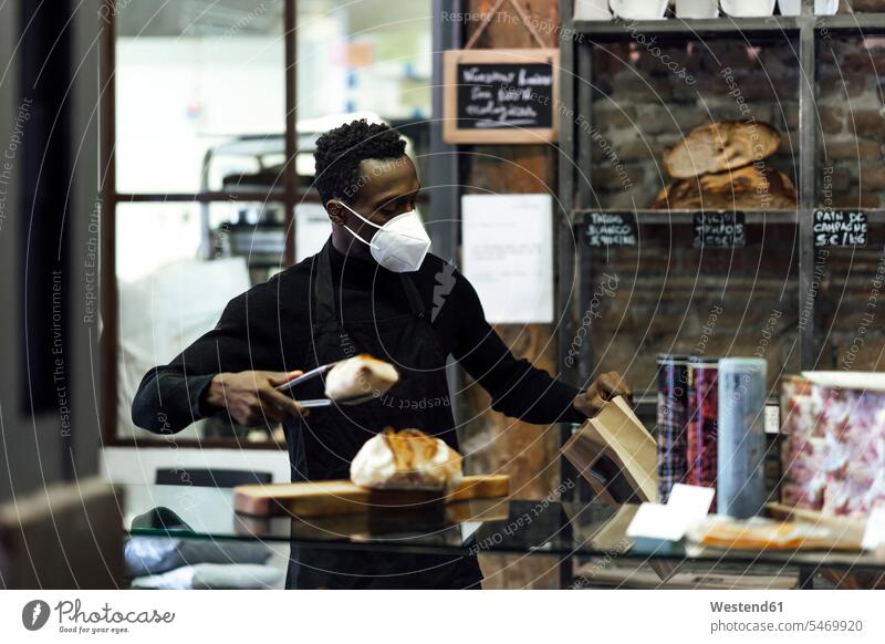 Verkäufer mit Gesichtsmaske beim Verpacken von Brot in einer Bäckerei stehend Farbaufnahme Farbe Farbfoto Farbphoto Außenaufnahme außen draußen im Freien Tag