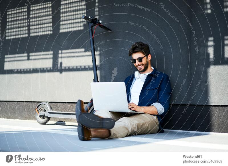 Lächelnder junger Mann sitzt auf dem Boden neben seinem Elektroroller mit Laptop sitzen sitzend benutzen Männer männlich daneben E-Scooter Notebook Laptops