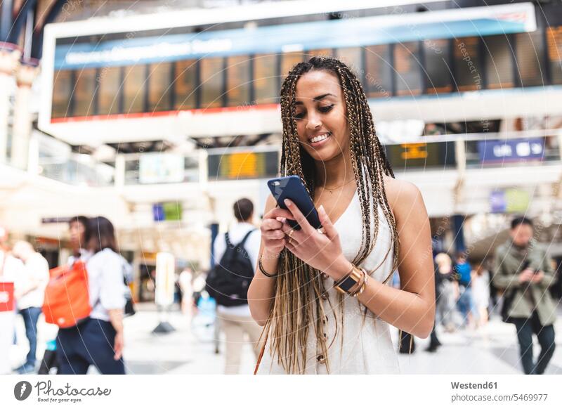 Porträt einer glücklichen jungen Frau am Bahnhof mit Smartphone, London, Großbritannien Leute Menschen People Person Personen gemischtrassig 1 Ein ein Mensch