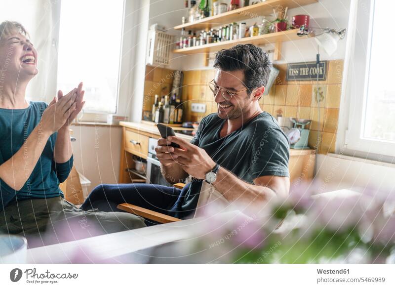 Glückliches Paar sitzt am Tisch in der Küche und benutzt ein Smartphone Informationen Telekommunikation telefonieren Handies Handys Mobiltelefon Mobiltelefone