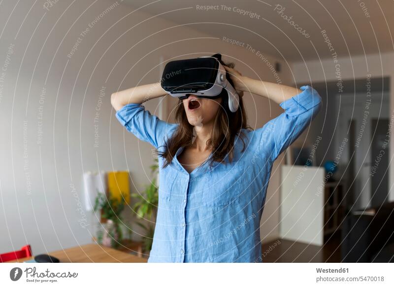 Junge Frau mit geöffnetem Mund schaut durch einen Virtual-Reality-Simulator, während sie im Home-Office steht Farbaufnahme Farbe Farbfoto Farbphoto Spanien