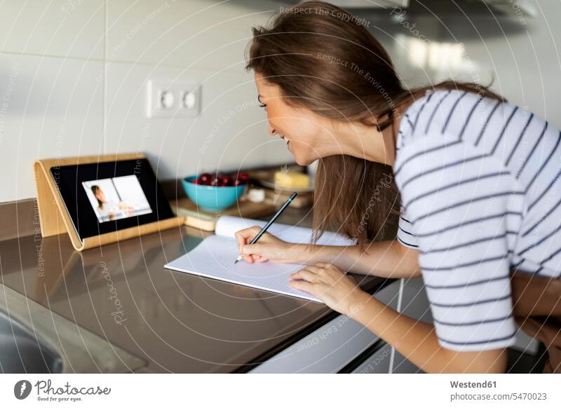 Junge Frau schaut sich zu Hause ein Video über eine digitale Tafel an und schreibt ein Rezept in ein Buch Farbaufnahme Farbe Farbfoto Farbphoto Spanien