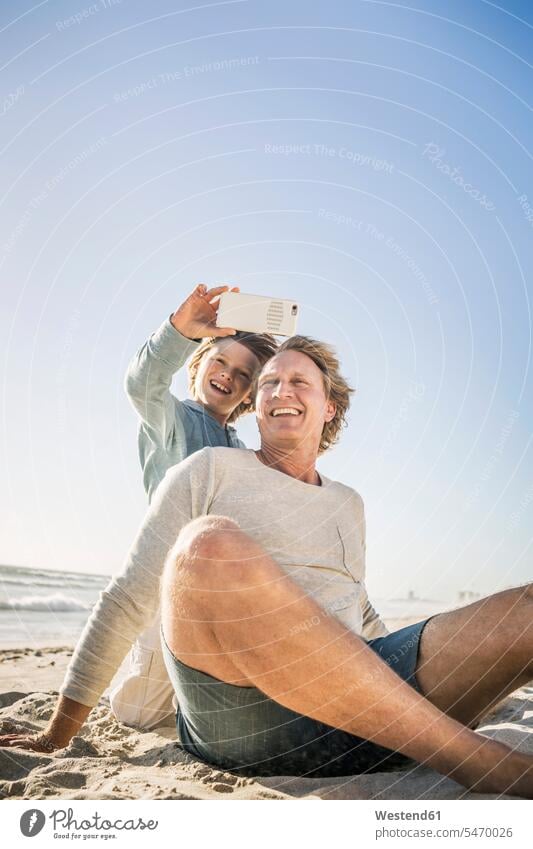 Vater amüsiert sich mit seinem Sohn am Strand und macht Smartphone-Fotos Leute Menschen People Person Personen Europäisch Kaukasier kaukasisch 2 2 Menschen