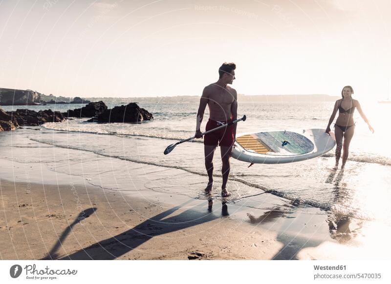 Frankreich, Bretagne, junges Paar mit einem SUP-Board am Meer zusammen Meere tragen transportieren Paddleboard Paddelbrett Gemeinsam Zusammen Miteinander