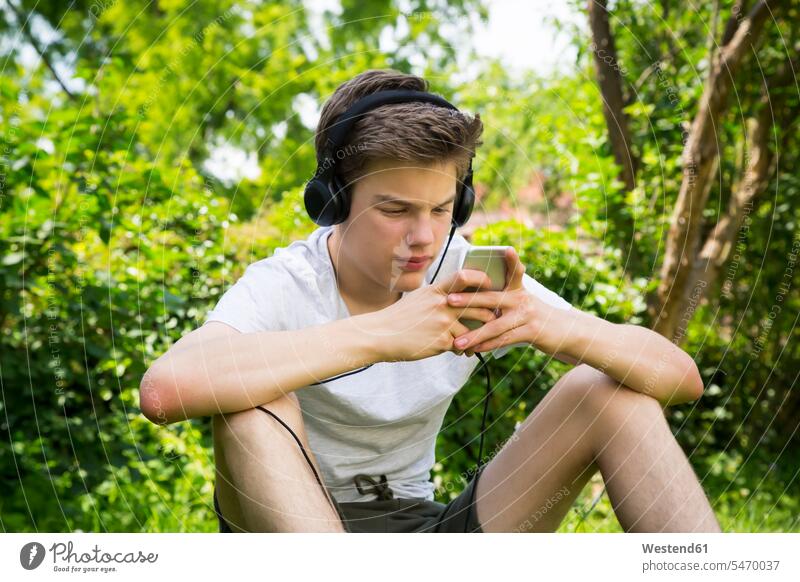 Junge sitzt im Garten und hört Musik mit Kopfhörern und Smartphone iPhone Smartphones Kopfhoerer hören hoeren Gärten Gaerten Buben Knabe Jungen Knaben männlich
