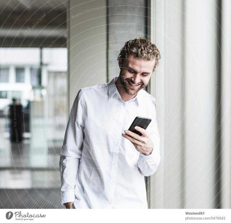 Lächelnder junger Geschäftsmann schaut auf Handy am Fenster im Büro Businessmann Businessmänner Geschäftsmänner lächeln Mann Männer männlich Mobiltelefon
