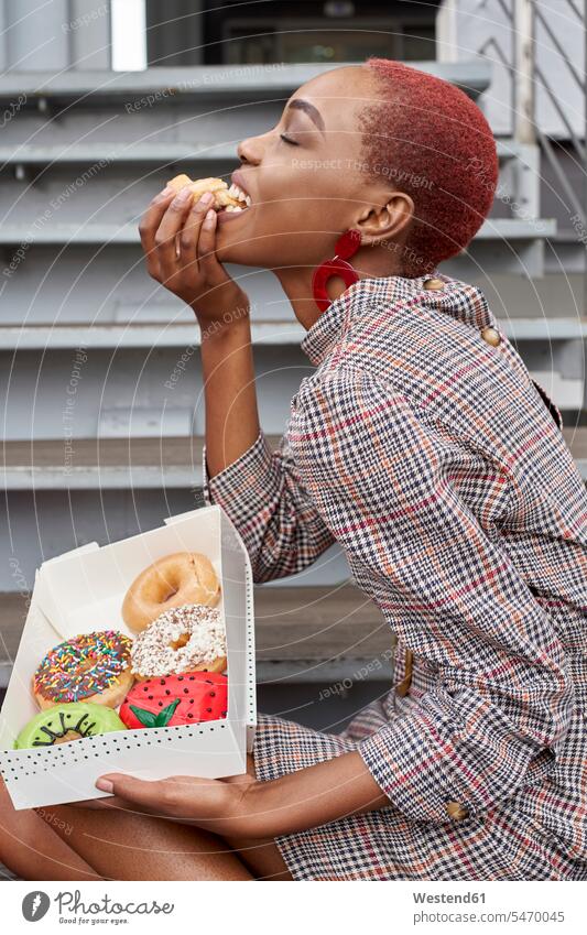 Junge Frau isst einen Doughnut aus der Schachtel Leute Menschen People Person Personen Afrikanisch Afrikanische Abstammung dunkelhäutig Farbige Farbiger