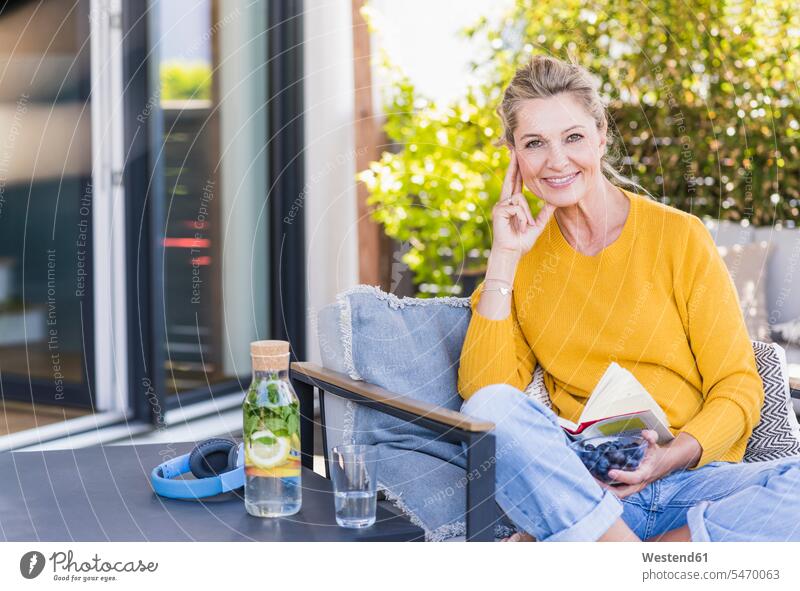 Porträt einer lächelnden reifen Frau, die auf einer Terrasse sitzt, mit einer Schale Blaubeeren und einem Buch Flaschen Glasflaschen Gläser Trinkglas