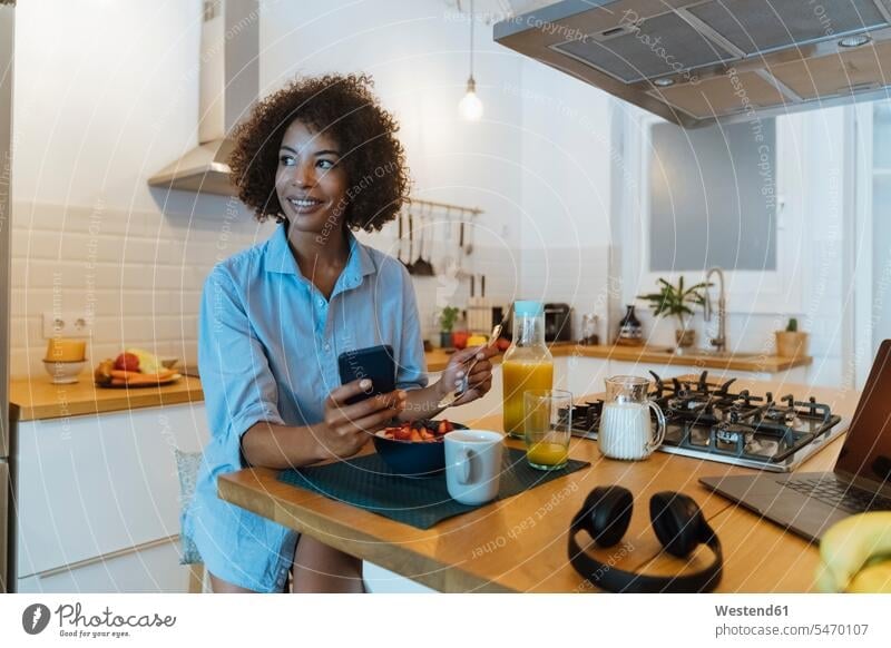 Frau frühstückt in ihrer Küche, mit Smartphone benutzen benützen lächeln Laptop Notebook Laptops Notebooks Küchen sitzen sitzend sitzt iPhone Smartphones