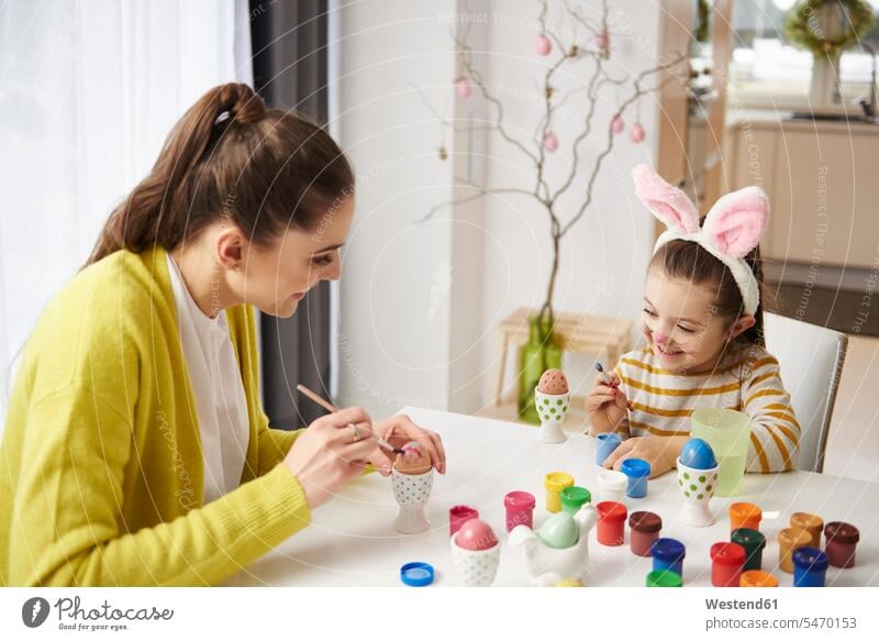 Glückliche Mutter und Tochter mit Hasenohren sitzen am Tisch und bemalen Ostereier Ostern Osterfest anmalen Häschenohren Mädchen weiblich glücklich
