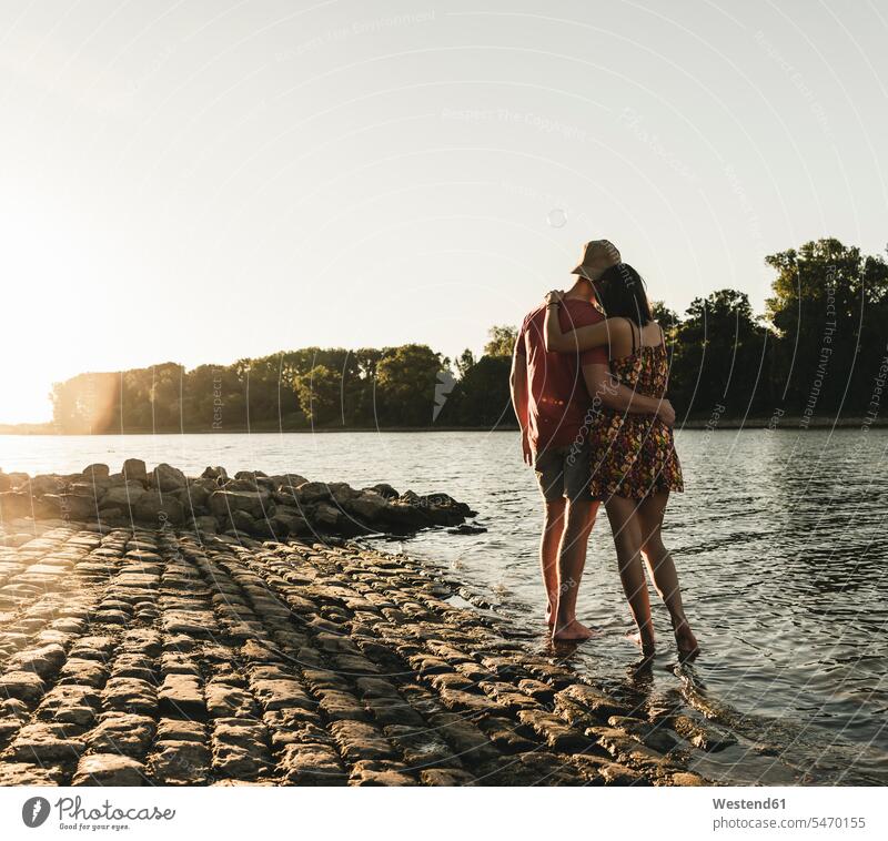 Rückansicht eines jungen Paares, das sich bei Sonnenuntergang an einem Fluss umarmt Sonnenuntergänge umarmen Umarmung Umarmungen Arm umlegen Fluesse Fluß Flüsse