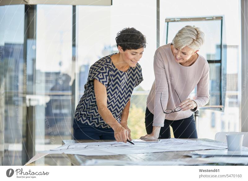 Zwei lächelnde Geschäftsfrauen besprechen einen Plan auf dem Schreibtisch im Büro Businesswomen Businessfrauen Businesswoman Office Büros Pläne arbeiten Arbeit