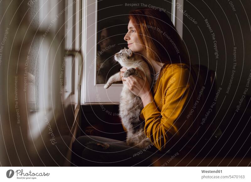 Lächelnde rothaarige Frau sitzt mit ihrer Katze am offenen Fenster zu Hause Leute Menschen People Person Personen Alleinstehende Alleinstehender Singles