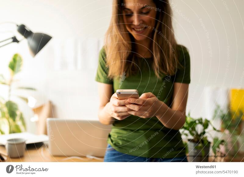 Lächelnde junge Frau, die ein Smartphone benutzt, während sie im Home-Office am Schreibtisch steht Farbaufnahme Farbe Farbfoto Farbphoto Spanien