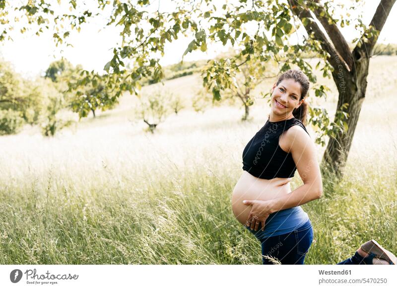 Junge schwangere Frau mit Babybauch, auf einer Wiese stehend Oberkoerper Oberkörper Torso Torsos Bäuche anfassen Berührung freuen Glück glücklich sein