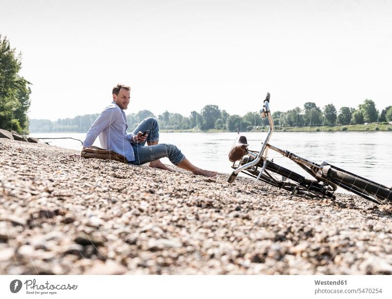Erwachsener Mann mit Fahrrad und Smartphone am Rheinufer sitzend Männer männlich Bikes Fahrräder Räder Rad sitzt Ufer Radfahrer Fahrradfahrer iPhone Smartphones