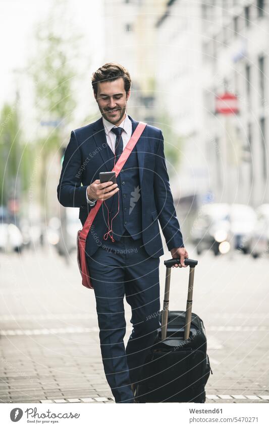 Lächelnde Geschäftsmann zu Fuß in der Stadt mit Handy und Koffer Deutschland Smartphone iPhone Smartphones Reisende Reisender Freiberufler freiberuflich