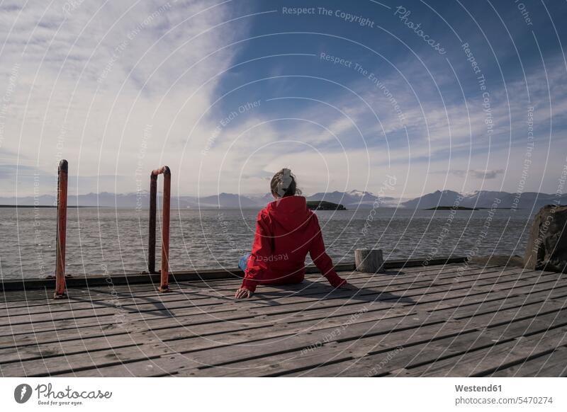 Junge Frau sitzt auf einem Steg und schaut auf das Meer, Südost-Island Jacken sitzend rote roter rotes frei auf Achse in Bewegung Abenteuer abenteuerlich Wolken