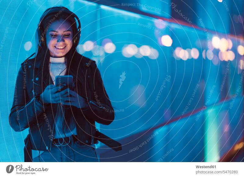 Porträt eines lächelnden Teenager-Mädchens mit Kopfhörern, das vor einer blauen Glasscheibe steht und auf ein Smartphone schaut Leute Menschen People Person