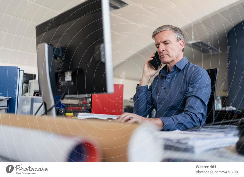 Hübscher männlicher Berufstätiger, der im Büro am Computerschreibtisch sitzt und mit dem Handy telefoniert Farbaufnahme Farbe Farbfoto Farbphoto Deutschland