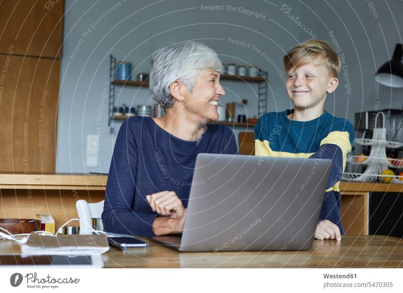 Lächelnder Enkel steht neben der Großmutter und arbeitet zu Hause am Laptop Farbaufnahme Farbe Farbfoto Farbphoto Innenaufnahme Innenaufnahmen innen drinnen Tag
