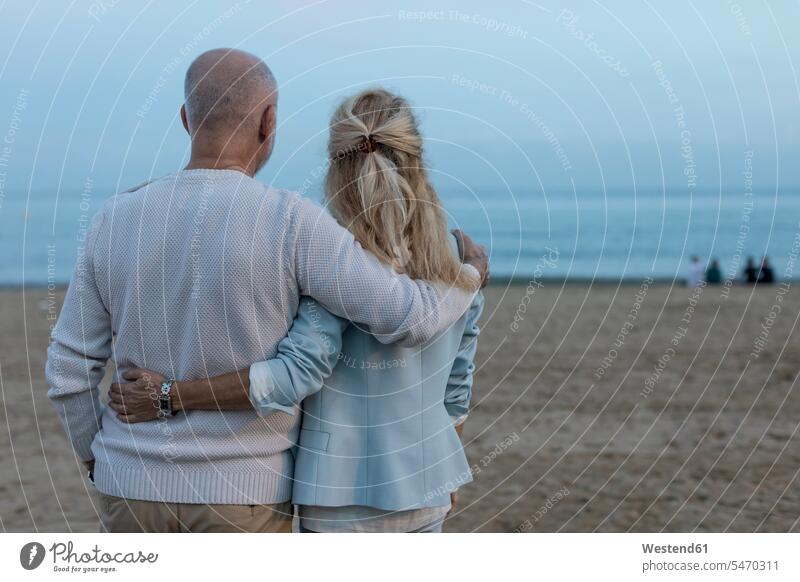 Spanien, Barcelona, Rückansicht eines sich umarmenden älteren Paares am Strand in der Abenddämmerung Umarmung Umarmungen Arm umlegen Seniorenpaar älteres Paar