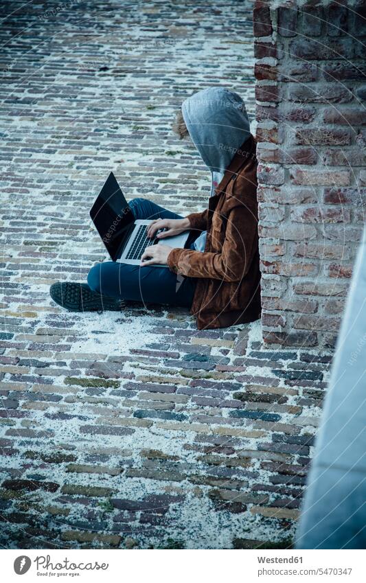 Teenager, der einen Laptop benutzt und auf einem Steinboden in der Stadt sitzt Kriminalitaet Kriminelle Kapuzen Rechner Laptops Notebook Notebooks sitzend Muße