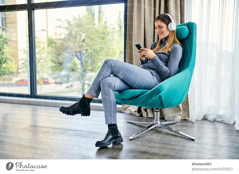 Frau sitzt im Sessel, hört Musik mit Kopfhörern, benutzt Smartphone Leute Menschen People Person Personen Europäisch Kaukasier kaukasisch erwachsen Erwachsene