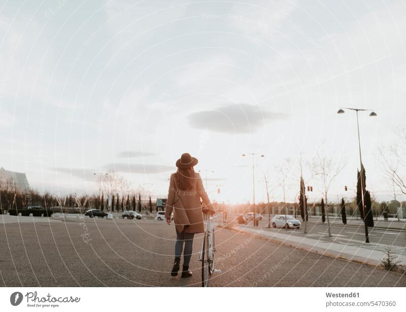 Junge Hipster Frau geht mit einem Fahrrad während des Sonnenuntergangs Hut Hüte Mantel Mäntel Jacke schieben anschieben Sanfte Mobilität Nachhaltige Mobilität