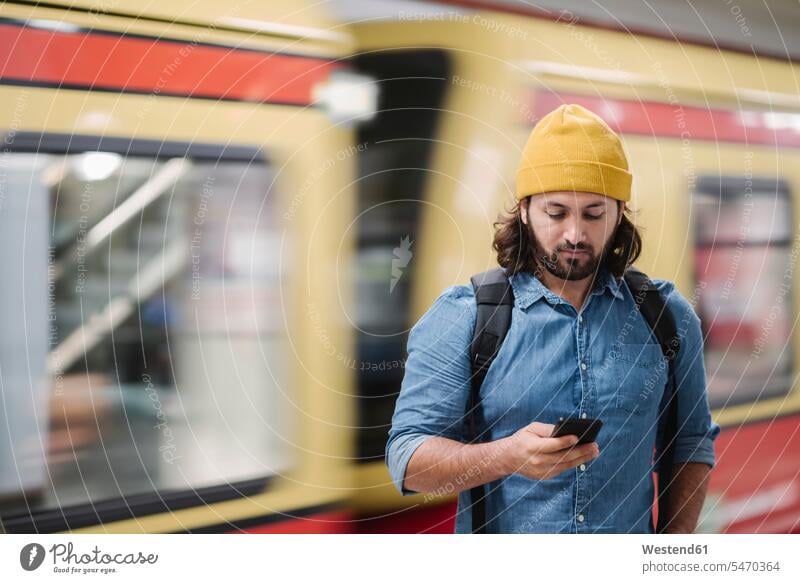 Porträt eines Mannes mit Rucksack, der am Bahnsteig auf ein Smartphone schaut, während er wartet, Berlin, Deutschland Leute Menschen People Person Personen