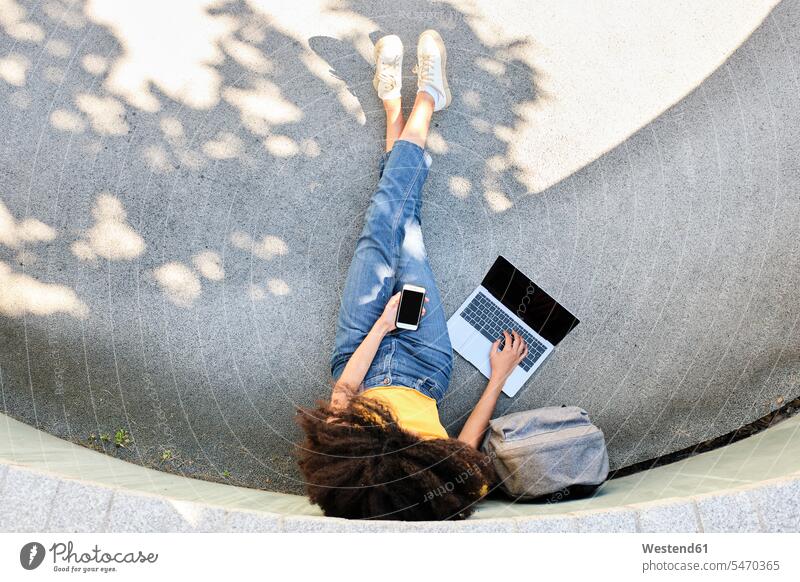 Student mit Mobiltelefon, der einen Laptop benutzt, während er auf dem Fußweg sitzt Farbaufnahme Farbe Farbfoto Farbphoto Außenaufnahme außen draußen im Freien