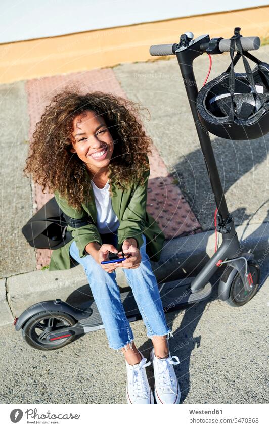 Glückliche junge Frau hält ein Smartphone in der Hand, während sie mit einem elektrischen Roller auf dem Bürgersteig sitzt Farbaufnahme Farbe Farbfoto Farbphoto