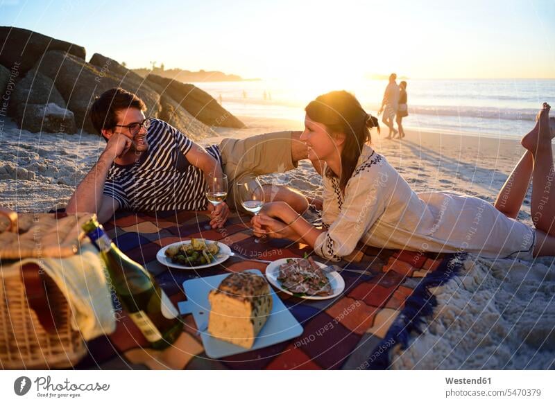 Glückliches Paar macht bei Sonnenuntergang ein Picknick am Strand Sonnenuntergänge Beach Straende Strände Beaches Pärchen Paare Partnerschaft picknicken