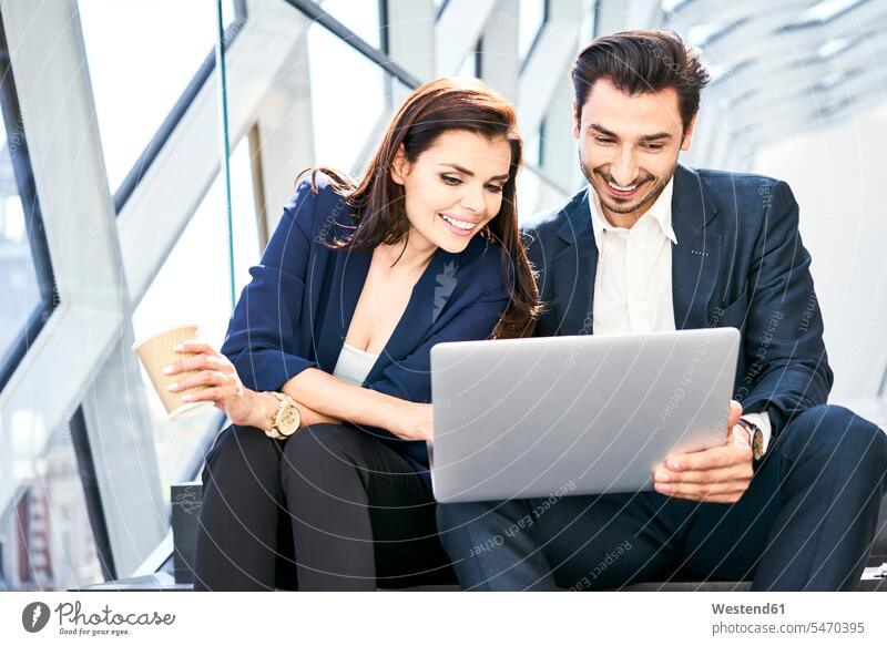 Lächelnde Geschäftsfrau und Geschäftsmann teilen sich Laptop auf der Treppe in einem modernen Büro Notebook Laptops Notebooks sitzen sitzend sitzt lächeln