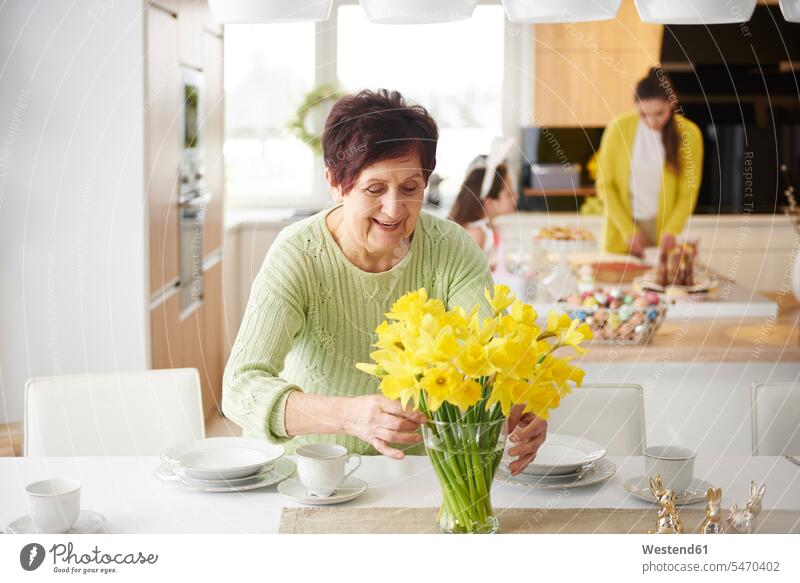 Ältere Frau arrangiert Blumen auf dem Esstisch mit der Familie im Hintergrund Mädchen weiblich Blüte anrichten Familien Zuhause zu Hause daheim Esstische