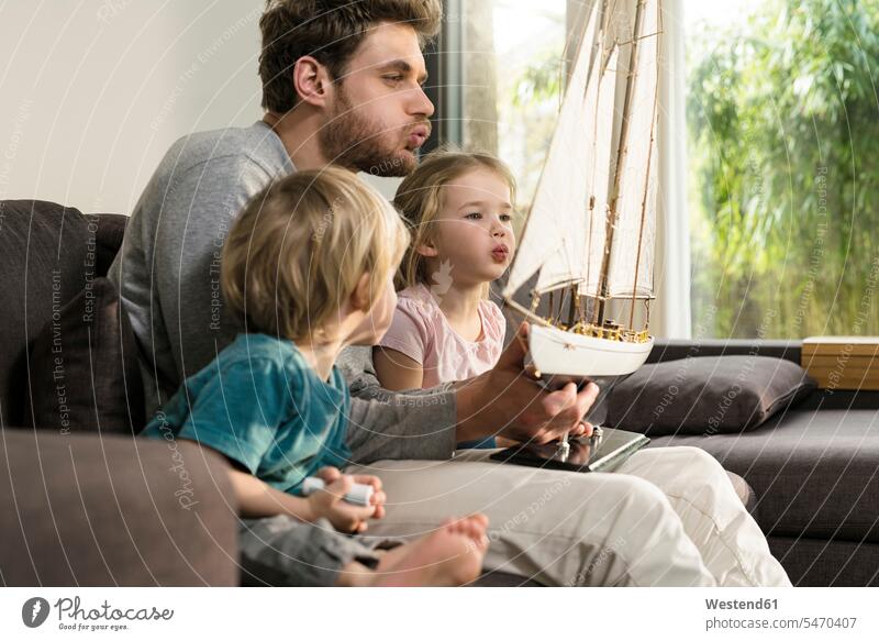 Vater und Kinder blasen zu Hause auf der Couch in die Segel eines Spielzeug-Modellschiffs Sofa Couches Liege Sofas pusten Papas Väter Vati Vatis Papis Zuhause