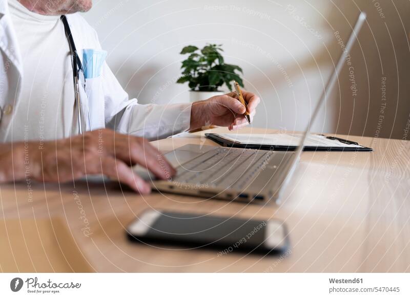 Arzt benutzt Laptop, während er in der Klinik am Schreibtisch sitzt Farbaufnahme Farbe Farbfoto Farbphoto Innenaufnahme Innenaufnahmen innen drinnen Tag