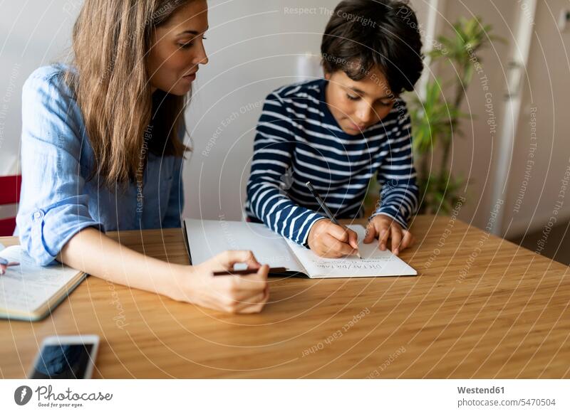 Weibliche Tutorin sieht den Jungen an, wie er Hausaufgaben in einem Buch auf dem Tisch schreibt Farbaufnahme Farbe Farbfoto Farbphoto Spanien Freizeitkleidung