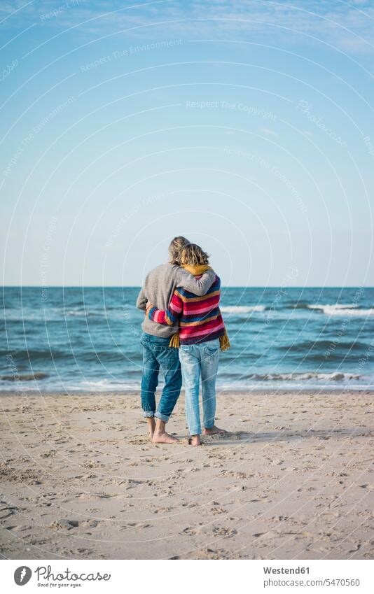 ture Paar, das am Strand steht und die Arme umeinander legt und auf das Meer schaut Meere Beach Straende Strände Beaches Pärchen Paare Partnerschaft stehen