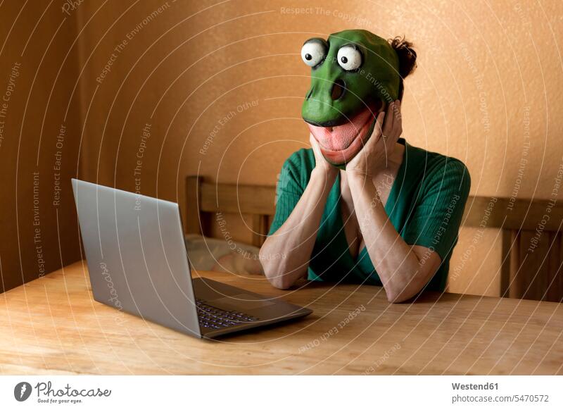 Verspielte reife Frau mit Froschmaske mit Laptop auf dem Tisch, die während der Ausgangssperre zu Hause sitzt Farbaufnahme Farbe Farbfoto Farbphoto Deutschland