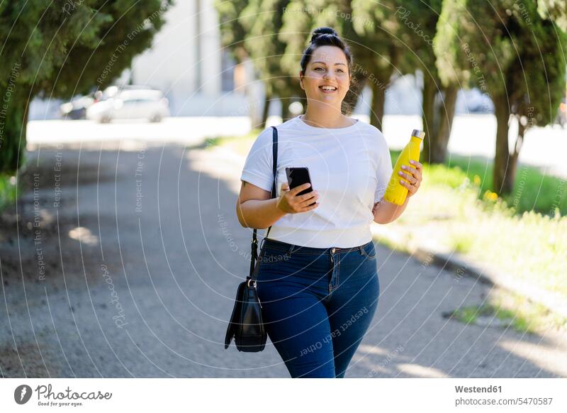 Lächelnde kurvenreiche junge Frau mit Handy und Flasche in einem öffentlichen Park Leute Menschen People Person Personen Europäisch Kaukasier kaukasisch 1 Ein