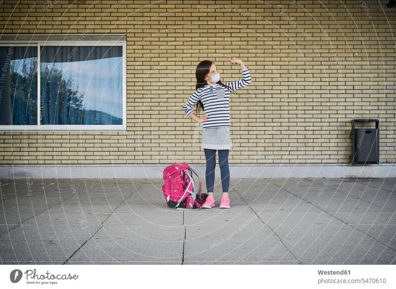 Mädchen mit Schultasche mit Maske vor dem Gebäude stehend und ihre Muskeln beugend Ausbildung Schueler Schulkinder Schüler steht Gesund kräftig stark Stärke