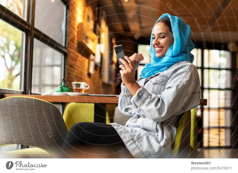 Junge Frau trägt einen türkisfarbenen Hijab und benutzt ein Smartphone in einem Cafe Kopftücher Telekommunikation telefonieren Handies Handys Mobiltelefon